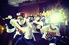 A close-up shot of a performance by four young men in a hip-hop dance crew dressed in black slacks, white t-shirts, white face masks, and red baseball caps.