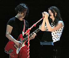 On the left, a man in red pants and a black T-shirt with black hair down to his chin holding a red guitar. On the right, a woman wearing a white shirt with black polka dots standing behind a red microphone stand.