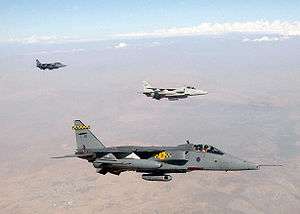 Grey jet aircraft flying above desert, with white aircraft further out, trailed be another jet.