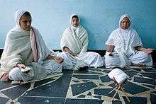 Three women in white saris, meditating cross-legged on a floor