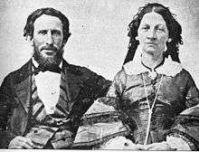 He has dark bushy hair and a beard and is wearing a three-piece suit with wade lapels and a bow tie. She has dark hair and wears a 19th-century dress with lace collar and bell sleeves.