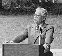 A man wearing a suit and glasses standing at a podium outside.