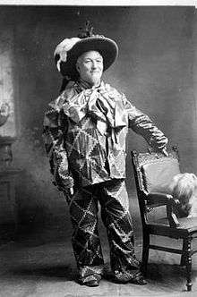 A man in a clown costume standing with his left arm on top of a leather chair.