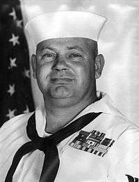A black and white image showing the head and upper torso of Williams wearing his military dress uniform with hat and ribbons. An American flag is visible in the background