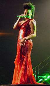 With long brown hair, a woman holds a microphone wearing a long red dress.