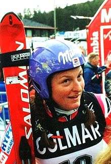 Janica Kostelić standing and holding skis, wearing a helmet and dressed in a ski racing suit.