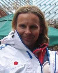 A smiling man with long dark blond hair and a piercing in his left eyebrow is wearing a white winter sports coat over a red jacket; the white coat has a white badge with a red circle in the center.