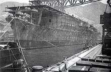 Photograph of a large ship floating in the water next to a long dock. The photograph is taken from a location on the dock in front of the ship's bow and looking back towards its stern. There are many cables and chains tying the ship to the dock. There is scaffolding all along the ship's side visible between the waterline and the ship's flat deck. A large metal structure runs horizontally from the dock to a location somewhat above the ship's upper deck. There are mountains visible in the distance behind the ship and the dock, and some sky is visible above the mountains.