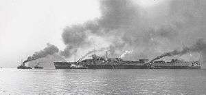 Small tugboats surround a partially completed large ship; dark smoke is rising from the boats
