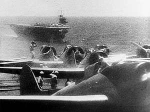 Photograph showing four aircraft lined up with several men working nearby. The sea and a complete ship are visible in the distance, along with the horizon and some sky.