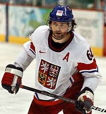 An ice hockey player holds his stick across his body. He is wearing a blue helmet and a blue and white uniform.