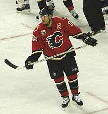 A hockey player in a red and black uniform with a stylized C logo on his chest skates across the ice.