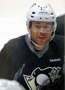 Upper body of a man looking to his right.  He is in a black jersey with a stylized penguin logo on his chest and is wearing a white helmet with a visor.