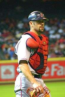 A baseball player walks off the field in his catcher's gear; he is wearing a chest protector, with a catcher's mask in hand