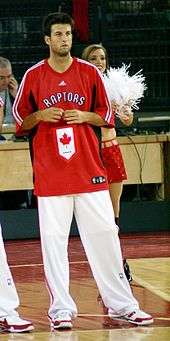A man, wearing white pants and a red t-shirt with the word "RAPTORS" on the front, is standing on a basketball court.