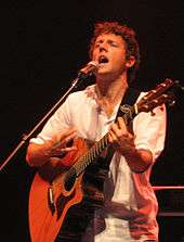 A curly haired man, strumming a guitar and wearing a white shirt.