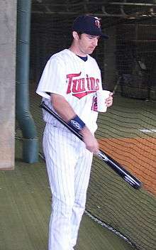 A man in a white pinstriped baseball uniform with "Twins" on the chest holds a baseball bat and a cup.