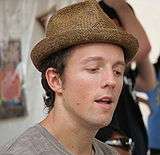 Jason Mraz wearing a brown hat and grey T-shirt, looking downwards