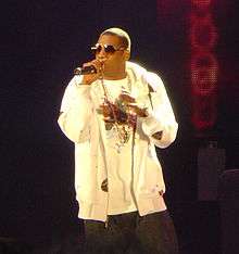 A image of an African American male wearing sunglasses with black pants and a white shirt and jacket. He is speaking into a microphone while holding it with his right hand. In the mostly dark background, a red colored wall with a design can be seen.