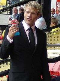 The head and torso of a man wearing a black dinner jacket, white shirt and black tie. He is holding a drinks bottle.
