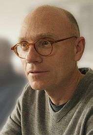 A bald man wearing glasses and a gray T-shirt looks off-screen.