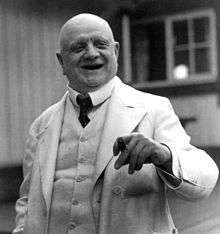 A jolly-looking older gentleman in a white suit, holding a cigar in his left hand and laughing