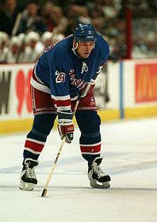 A Caucasian hockey player.  He wears a dark blue jersey and helmet.  He is bent over at the waist leaning on his stick.
