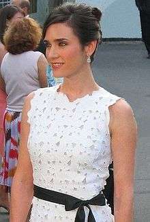 A brown haired woman, wearing a white dress with small flowers detail and a black ribbon in her hips. She is also wearing pendants. Behind her there is a woman dress with a white shirt and a white skirt with big flowers details.