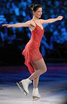 A female figure skater wearing a red dress, in profile as she lands a jump, extending her arms away from her perpendicular to the ice sheet for balance