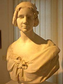 Photograph of a stone bust of a young woman looking directly at the reader. The stone appears to be white marble. Her hair is cut well above her shoulders. She is wearing a dress that hangs loosely from her shoulders, leaving her neck and upper chest bare.