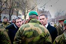 Two gray haired older men talk with a soldier wearing camouflage and a green beret who is facing away.