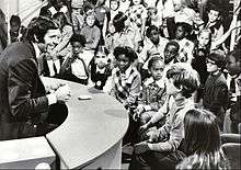 Black & white photo of a dark-haired Caucasian man seated in front of an audience of children.