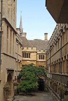 A narrow courtyard, longer than it is wide; a tree at the far end and hanging baskets of flowers on each side; tall stone buildings on each side, the ones at the far end topped with crenellations; a large archway on the left; above roof-level, a tall metal spire