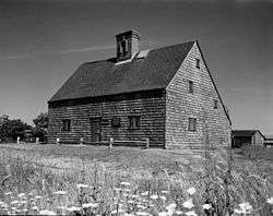 This black and white photo shows a saltbox-style house with split-rail fencing in front. A meadow with flowers is in the foreground, and small buildings associated with the house are in the background.