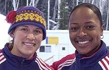 Two women, both smiling.  The one on the left is Jill Bakken, and the one on the right is Vonetta Flowers.  Bakken is wearing a winter hat, both are wearing U.S. team jackets.