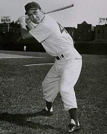 A man in a white baseball uniform holding his bat behind his head prepares to swing.