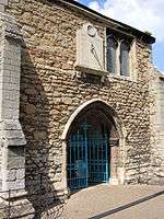 A two-storey medieval church porch with blue gates. A small square perpendicular window, offset to the right, is on the second level.