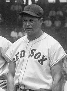 A man is pictured from his belt up looking to the left of the camera. His button-down baseball jersey says "RED SOX" across it and he is wearing a baseball cap.