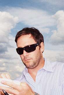 A man in his thirties, wearing black sunglasses, and a striped shirt. He is signing his autograph onto a piece of paper.