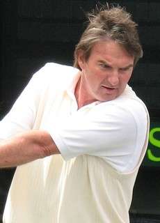 A brown-haired man dressed in a white shirt swings a two-handed backhand