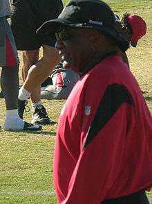 Candid waist-up photograph of Raye from the side standing on a football field, wearing a red and black pullover, a wide-brimmed hat bearing a Reebok logo and sunglasses