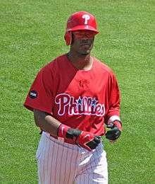 A dark-skinned man in a red baseball jersey and red left-handed batting helmet walks on a baseball field; he appears to be in his mid-twenties. His jersey reads "Phillies" in white and red script, with two blue starts dotting the "i"s.
