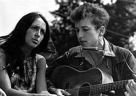Dylan is seated, singing and playing guitar. Seated to his right is a woman gazing upwards and singing with him.