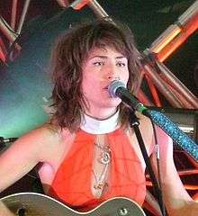 Head-and-shoulders colour photograph of Joan Wasser singing live in 2006. She is playing a guitar and singing into a microphone.