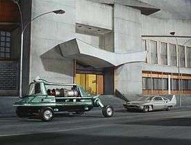 Two very different vehicles are parked at the side of a street in front of the entrance to a grey concrete building. The vehicle on the left is viridian green and of an eccentric design, with a turbine engine positioned behind a cockpit to seat the driver and passengers. The vehicle on the right is a car of a more standard appearance and grey in colour, although it is fitted with tail fins at the rear.