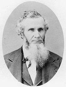 Formal portrait showing the head and shoulders of a white-haired, full-bearded man wearing a dark suit