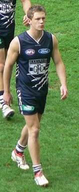 Wearing a navy sleeveless jersey and shorts, young male athlete strides from the grass surface of the playing arena.