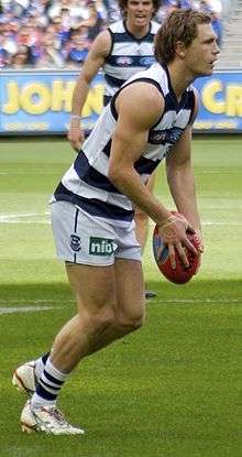 Young male athlete stands in a white and navy blue and striped sleeveless shirt and white shorts. He stands alone and looks to the right while preparing the kick the football.