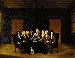 A painting showing eight bewigged men, sat around a table with papers and quills