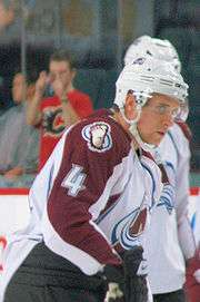 An ice hockey player stands facing the right of the camera. He is wearing a white helmet and a burgandy and white uniform with a large burgandy A on his chest.